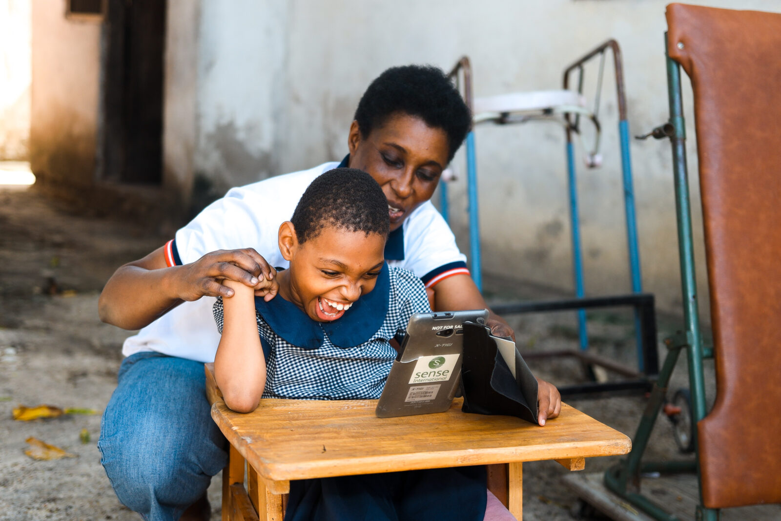 Young black girl with deafblindness, smiling ,  sat down with a tablet on a table. He mum is behind her , holding her right hand.