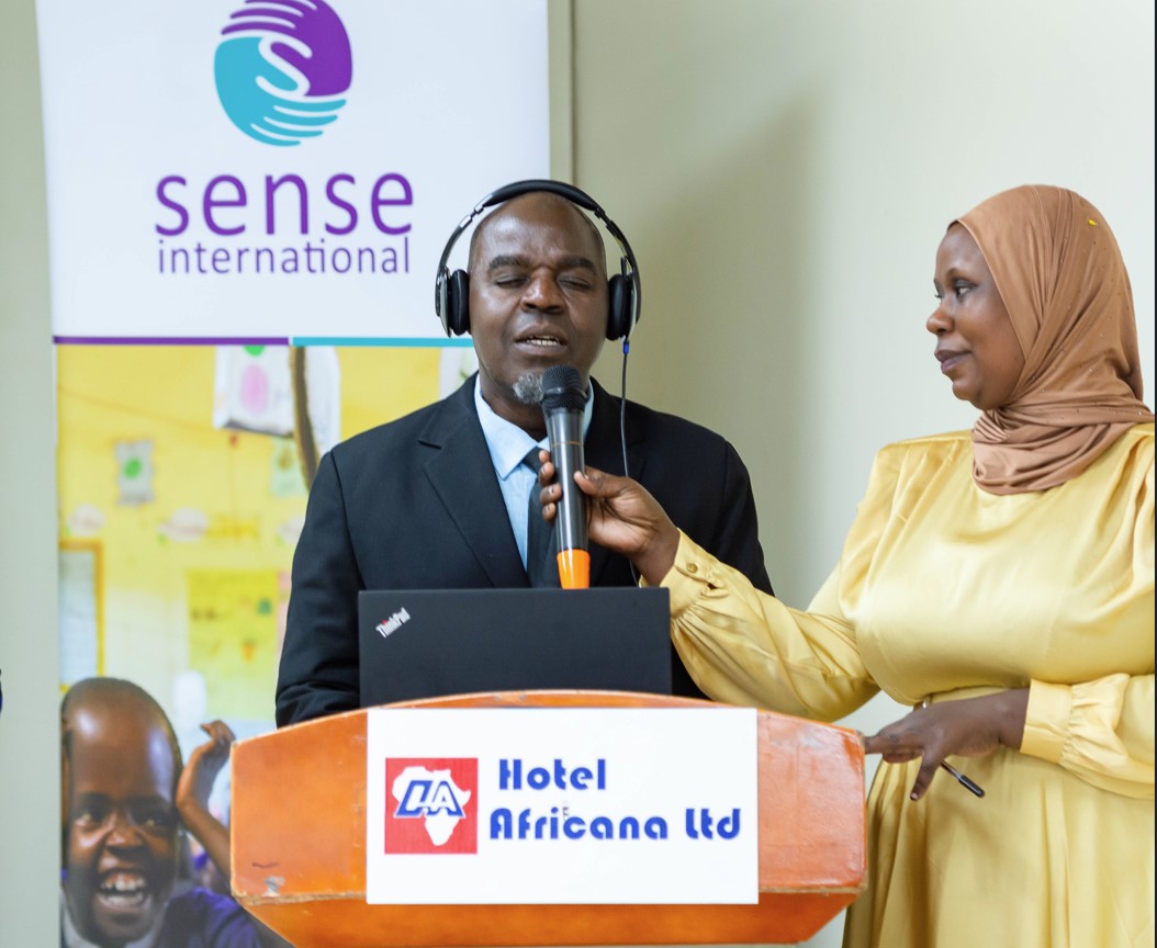 A black middle aged man with visual impairment wearing headphones. He is wearing a navy blue suit and stood in front of an audience giving a speech into a microphone which is being held by a middle aged black woman wearing a headscarf and yellow dress. A black middle aged man with visual impairment wearing headphones. He is wearing a navy blue suit and stood in front of an audience giving a speech into a microphone which is being held by a middle aged black woman wearing a headscarf and yellow dress.