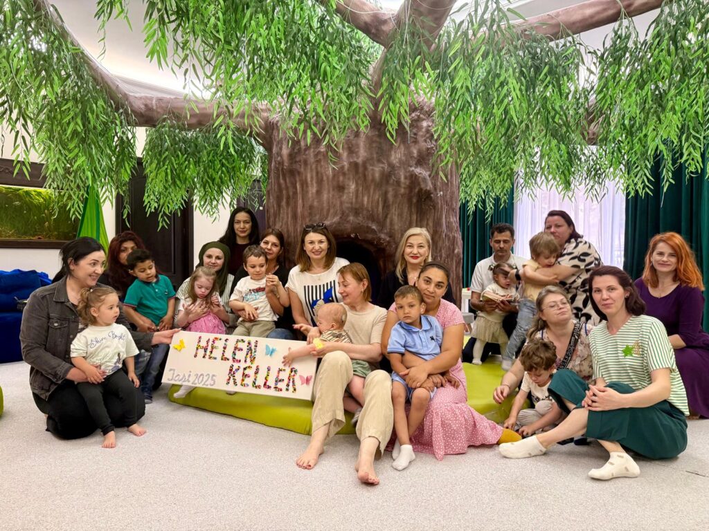 : A group of adults and children gathered in a room decorated with a large artificial tree, sitting together on the floor in front of the tree. Two of the adults hold a colorful sign that reads "Helen Keller – Iași 2025".