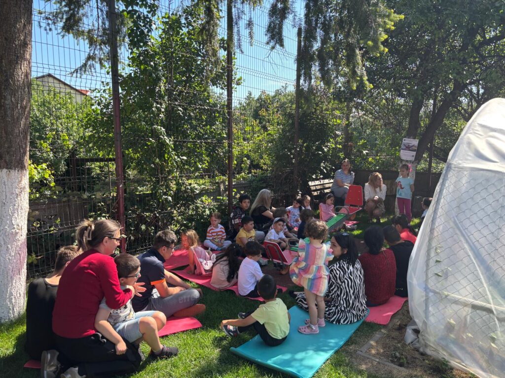 A group of young children and adults sit on colorful mattresses, on the grass, in a green courtyard, shaded by trees. The children are gathered in a semicircle, in a quiet setting of outdoor activity.