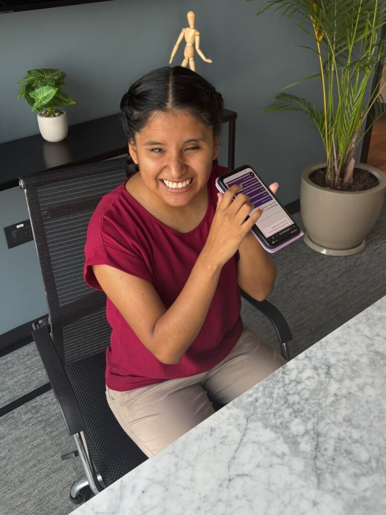 Foto de Milagro sentada en un escritorio escuchando un recurso en el Hub usando la aplicación Talkback en su teléfono móvil.