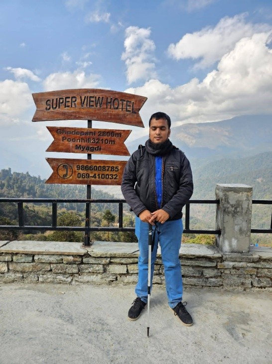 Jubayer stands in front of a wooden sign for Super View Hotel, which lists distances to Ghorepani and Poonhill, and phone numbers. He is holding  holds a white cane for visually impaired people and is dressed in a dark jacket, blue shirt, and blue jeans. The background features a mountainous landscape under a cloudy sky.